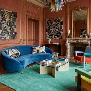 May include: A luxurious living room featuring a teal rug, a curved blue velvet sofa with patterned pillows, and a glass-top coffee table. The room has coral-colored walls, gold trim, and a colorful glass chandelier. Artwork and a fireplace complete the elegant space.