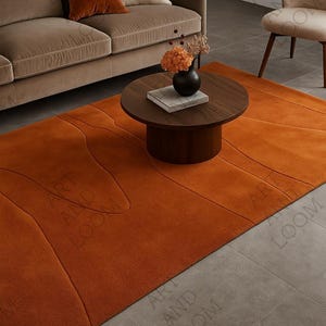 Peut inclure: Un tapis orange avec un motif abstrait en relief. Le tapis est dans un salon avec un canapé marron, des coussins, une table basse ronde en bois et une chaise. Les dimensions du tapis ne sont pas visibles.