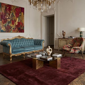 May include: An elegant living room featuring a teal velvet sofa with gold trim, a patterned armchair, and a burgundy rug. A glass-topped coffee table sits on a brass base, and a gold chandelier hangs from the ceiling.