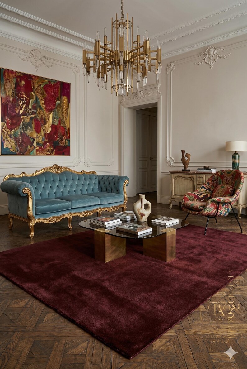 May include: An elegant living room featuring a teal velvet sofa with gold trim, a patterned armchair, and a burgundy rug. A glass-topped coffee table sits on a brass base, and a gold chandelier hangs from the ceiling.