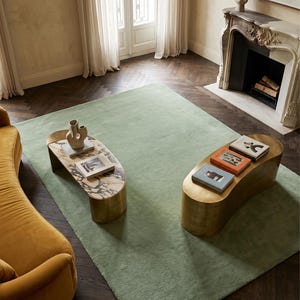 May include: An interior shot featuring a light green rug, two unique coffee tables, and a mustard-yellow sofa. The coffee tables have a gold base and marble or stone tops. Books and decorative vases are on the tables. The room has a fireplace and large windows.