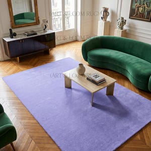 May include: A large, rectangular, solid lavender rug dominates a room with hardwood floors. A green curved sofa and a wooden coffee table with a vase and books are also in the room. The room has a mirror and artwork.