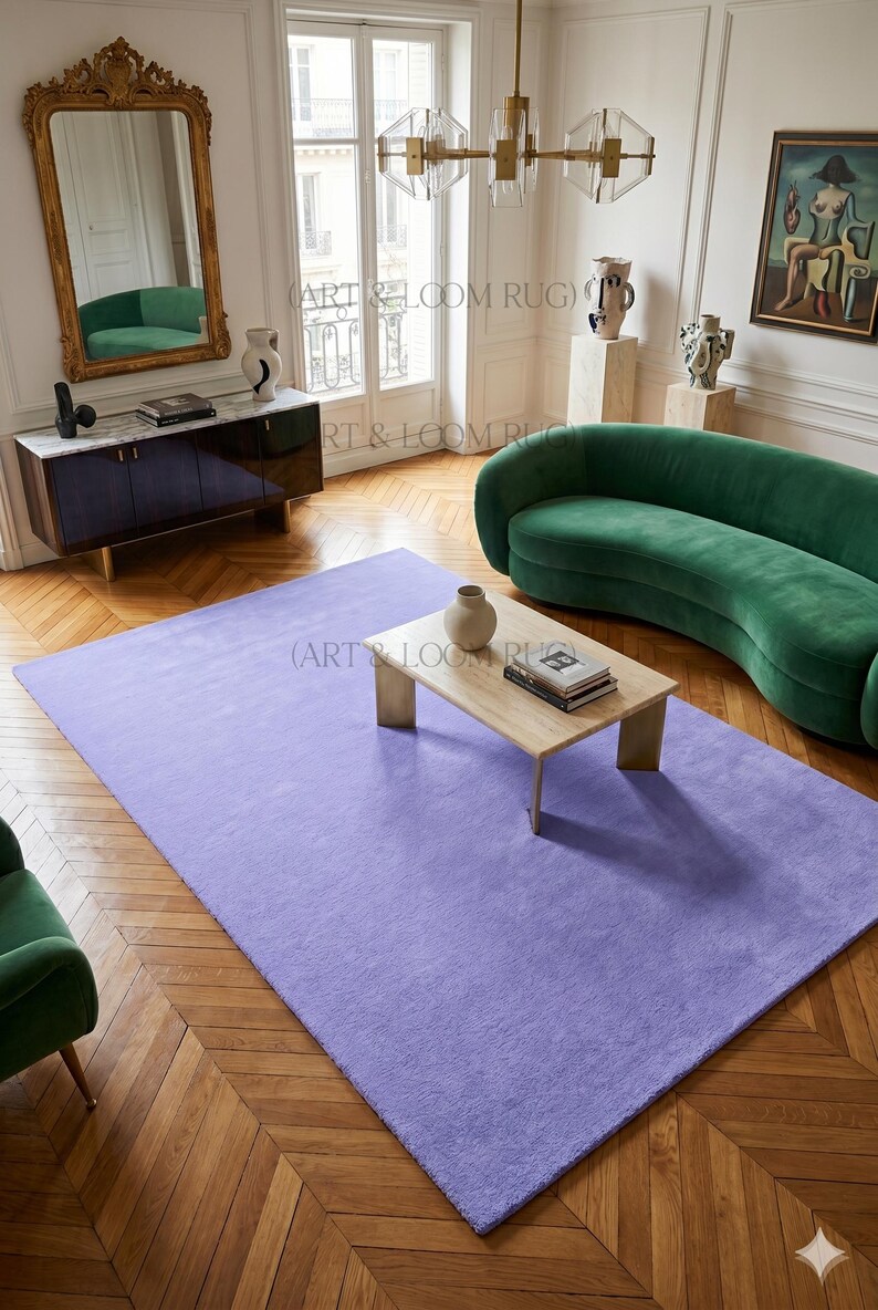 May include: A large, rectangular, solid lavender rug dominates a room with hardwood floors. A green curved sofa and a wooden coffee table with a vase and books are also in the room. The room has a mirror and artwork.