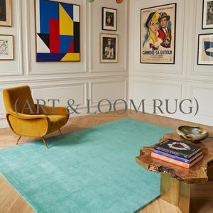 May include: A teal-colored rug dominates the room, complemented by a mustard-yellow armchair. The room features artwork, including a colorful abstract painting and a vintage movie poster. A wooden coffee table displays books with the text "ART DECO JEWELS" and "MID-CENTURY MASTERS". The text "(ART & LOOM RUG)" is visible.