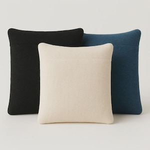 Op de afbeelding: Drie vierkante decoratieve kussens in zwart, crème en marineblauw. De kussens hebben een eenvoudig, minimalistisch ontwerp met een textuurstof. De kussens zijn in een groep gerangschikt en tonen hun verschillende kleuren en vormen.