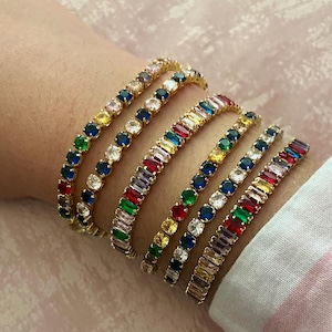 Op de afbeelding: Een verzameling goudkleurige armbanden, elk versierd met een levendige reeks kleurrijke edelstenen. De armbanden hebben een verscheidenheid aan vormen en maten van edelstenen, waaronder ronde en rechthoekige slijpsels, waardoor een oogverblindende weergave van kleur en glans ontstaat.