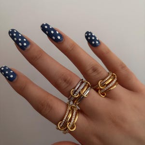 Puede incluir: Una mano que muestra tres anillos. Dos anillos son dorados con piedras transparentes, y uno es plateado y dorado. Las uñas están pintadas de azul marino con lunares blancos. Los anillos son un accesorio de joyería.