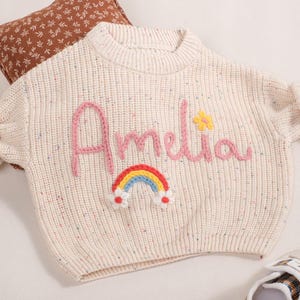 May include: Cream-colored knit sweater with the name "Amelia" embroidered in pink, a yellow flower, and a rainbow. The sweater has a speckled design and ribbed cuffs. A brown floral pillow is in the background.