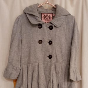 May include: A light gray double-breasted coat featuring a wide collar, five dark brown buttons, and three-quarter length sleeves with ruffled cuffs. The garment has a pleated waistline, two side pockets, and a Juicy Couture label at the neckline.