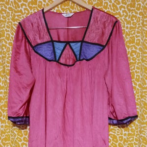 May include: A vintage pink satin dress with a square neckline and three-quarter sleeves. The neckline features a geometric design with black, blue, and purple accents. The dress is hanging on a wooden hanger against a yellow patterned background.