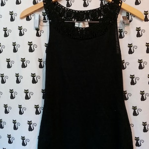 May include: A black tank top with a crocheted neckline hangs on a wooden hanger. The top is sleeveless and has a simple, elegant design. The background features a repeating pattern of black cat silhouettes.