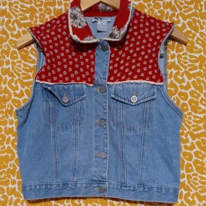 May include: A light blue denim vest with a red floral patterned collar and shoulder detail. The collar features a white floral design. The vest has two front pockets and silver buttons. The vest is hanging on a wooden hanger.
