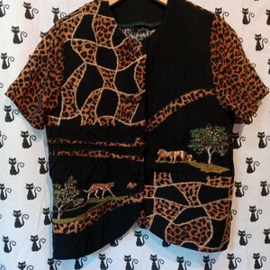 Puede incluir: Un chaleco negro y marrón con estampado de animales y mangas cortas. El chaleco presenta escenas bordadas de animales y árboles. Las mangas y secciones del chaleco tienen un estampado de leopardo. El chaleco está colgado de una percha de madera.