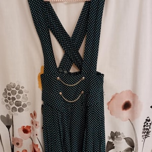 May include: A dark green polka dot overall dress with a pleated skirt. The dress features gold-colored buttons and chain detailing on the bodice. The straps cross in the back and are held up by a pink beaded hanger.