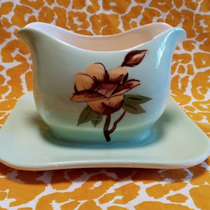 Puede incluir: Una salsera azul claro vintage con un platillo cuadrado a juego. La salsera presenta un diseño floral con acentos marrones y verdes. El interior del recipiente es de color crema.