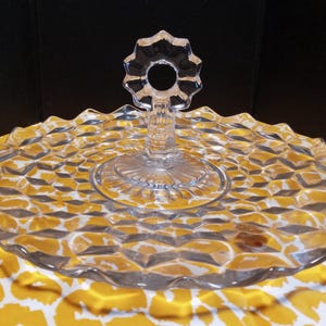 May include: Clear glass serving tray with a diamond pattern and a decorative handle. The tray has a scalloped edge and is designed for serving appetizers or desserts. The handle is shaped like a flower.