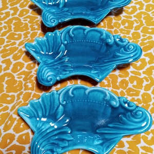 May include: Three turquoise ceramic decorative dishes with ornate, shell-like designs. Each dish features a scalloped edge and a detailed, swirling pattern. The dishes are arranged on a yellow and white patterned surface.