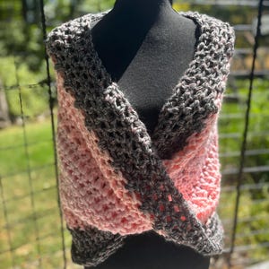 May include: A handmade, open-weave crochet wrap in shades of pink and gray. The wrap is draped over a black mannequin, showcasing its design and texture. The yarn appears soft and chunky, perfect for cooler weather.