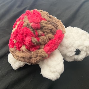 May include: A handmade crochet turtle plushie. The turtle has a shell with red, brown, and tan sections. The head, feet, and tail are white. The turtle has a black eye.