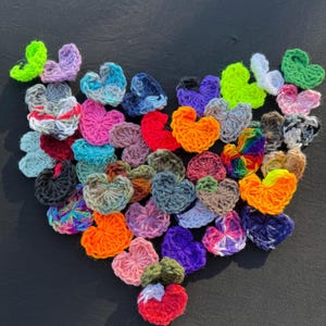 May include: A collection of crocheted hearts in various colors, arranged in a heart shape. The hearts are made of yarn and include shades of red, orange, yellow, green, blue, purple, and pink. The background is a dark, solid color.