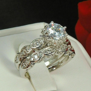 Peut inclure: Un ensemble de bagues de mariage argentées. La bague de fiançailles présente une grosse pierre précieuse ronde et transparente entourée de petites pierres. L'alliance a un design délicat en forme de feuille avec des pierres incrustées.