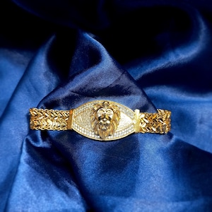 May include: A gold-colored bracelet with a lion's head centerpiece. The lion's head is surrounded by small, clear stones. The bracelet has a woven chain design on each side and is set against a blue background.