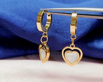 May include: Gold-colored hoop earrings with a heart-shaped pendant. The pendant features a white, heart-shaped inlay within a gold outline. The earrings are displayed against a blue background.