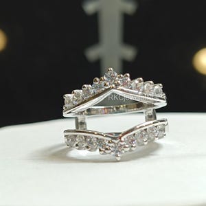 Könnte beinhalten: Ein silberner Ringschutz mit V-förmigem Design, besetzt mit mehreren kleinen, klaren Edelsteinen. Der Ringschutz ist so konzipiert, dass er um einen anderen Ring herum sitzt. Der Ring befindet sich auf einer weißen Oberfläche.