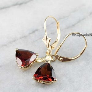 Peut inclure: Boucles d'oreilles dorées à fermoir à levier avec des pierres précieuses triangulaires rouge foncé. Les boucles d'oreilles ont un design classique avec une finition polie. Le texte "RKJewelsINC" est visible.