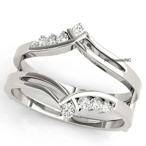 Puede incluir: Un protector de anillo plateado con un diseño curvo. El protector de anillo presenta una fila de pequeñas piedras preciosas transparentes. El diseño es elegante y moderno, adecuado para realzar un anillo de compromiso solitario.