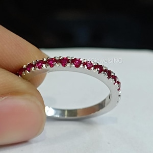 Puede incluir: Un anillo plateado con una banda de pequeñas gemas rojas vibrantes. Las gemas están engastadas muy juntas, creando una línea continua de color alrededor del anillo. El anillo se sostiene entre los dedos, mostrando su diseño.
