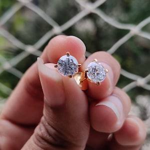 May include: A pair of gold stud earrings, each featuring a large, round, clear gemstone. The earrings are held up against a blurred green background, showcasing their sparkle and design.