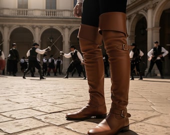 May include: Brown leather knee-high boots with a buckle closure. The boots are worn by a person in a historical setting, with other people in period clothing practicing sword fighting in the background.