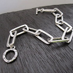 May include: A silver bracelet with a textured, rectangular chain and a round, textured charm.