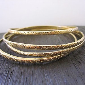 May include: Four gold-colored bangles with a textured crisscross pattern. The bracelets are stacked on top of each other, with the light reflecting off the polished metal. The bangles are a simple, elegant accessory.