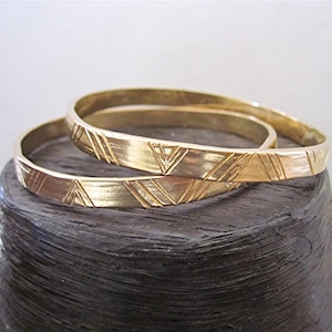 May include: Two gold bangles with a geometric pattern. The bangles are stacked on top of each other.