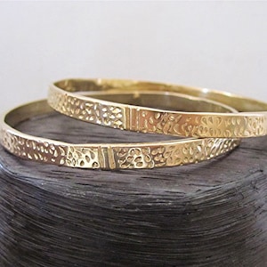 May include: Two gold bangles with a textured floral design. The bangles are stacked on top of each other.
