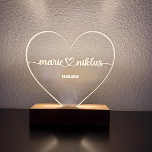 Könnte beinhalten: Eine herzförmige LED-Lampe mit den Namen "maric" und "niklas" sowie dem Datum "19.05.2018" in klarem Acryl eingraviert. Die Lampe steht auf einem rechteckigen Holzsockel und wird von einem warmen Licht erhellt.