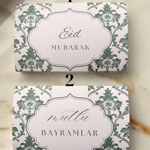 Könnte beinhalten: Zwei rechteckige Geschenkboxen mit einem grün-weißen floralen Design. Auf der oberen Box steht "Eid MUBARAK" in eleganter Schrift. Auf der unteren Box steht "mutlu BAYRAMLAR".