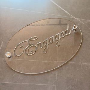Op de afbeelding: Een ovaal bord van doorzichtig acryl met het woord Engaged in een elegant cursief lettertype. Het bord is aan de linker- en rechterkant bevestigd met twee kleine, gefacetteerde, kristalachtige decoratieve knoppen voor een strakke uitstraling.