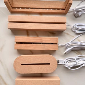 Könnte beinhalten: Eine Sammlung unfertiger Holz-Präsentationsständer mit LED-Leuchten und USB-Kabeln. Die Ständer haben verschiedene Formen, darunter rechteckig und oval, und sind zum Halten und Beleuchten von Objekten konzipiert. Das Holz hat eine natürliche, helle Farbe.