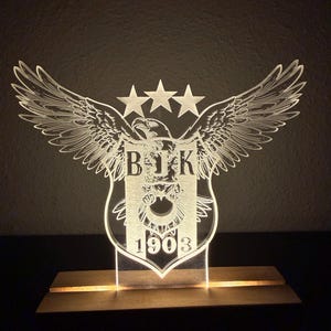 Peut inclure: Un emblème lumineux en acrylique, représentant un écusson avec un aigle, des ailes et trois étoiles. Les lettres "BJK" et le chiffre "1903" sont visibles. L'emblème est monté sur une base en bois, créant une lumière décorative.