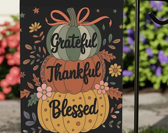 May include: Black garden flag with a stacked pumpkin design. The pumpkins are green, orange, and yellow, with the words "Grateful, Thankful, Blessed" in a decorative font. The flag is displayed on a black metal stand in a grassy yard.