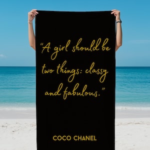 May include: Black beach towel with gold text that reads "A girl should be two things: classy and fabulous." The towel also includes the name "COCO CHANEL" in gold. The towel is held up on a beach with the ocean in the background.