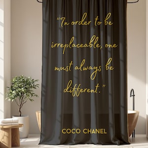 May include: A dark gray shower curtain with gold text that reads "In order to be irreplaceable, one must always be different." The text "COCO CHANEL" is below the quote. The curtain hangs in a bathroom with a wooden bathtub.