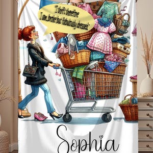 May include: White fleece blanket featuring a cartoon illustration of a woman pushing a shopping cart overflowing with clothes and accessories. The text "I thrift therefore I am broke but fabulously dressed" is in a speech bubble. The name "Sophia" is written at the bottom.