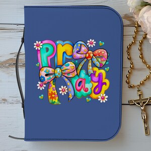 May include: A navy blue Bible cover with the word "PRAY" in colorful bubble letters and decorative bows. The cover has a black leather strap and is next to a gold rosary with a crucifix.