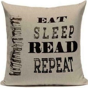 May include: Beige square pillow with the words "EAT SLEEP READ REPEAT" in black block letters. A stack of books is printed on the left side. The pillow is a decorative item for a reading nook or bedroom.