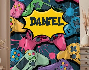 May include: A navy blue blanket featuring a colorful pattern of video game controllers in pink, green, yellow, and blue. The name "DANIEL" is displayed in a yellow speech bubble. The blanket has a splatter paint design.