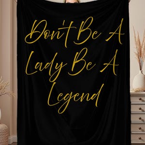May include: Black blanket with gold script text that reads "Don't Be A Lady Be A Legend". The blanket is held up by a person, and the background includes a beige wall, a white vase, and a decorative plant.